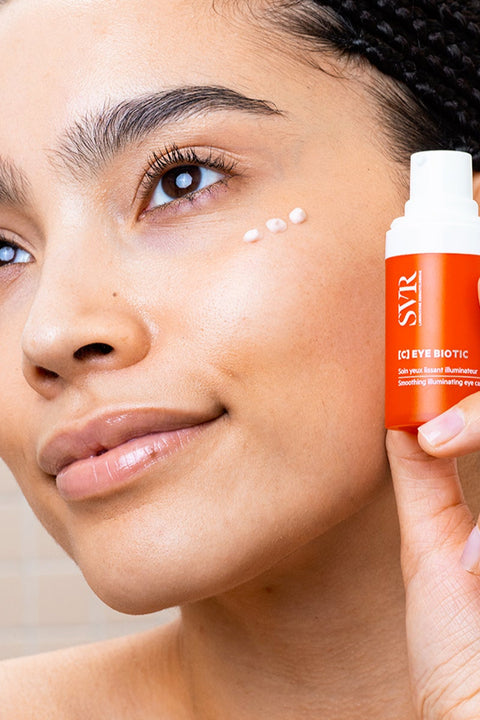 Woman applying brightening eye cream with a bottle of SVR [C] Eye Biotic in hand.